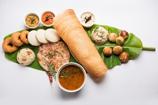 Group of South Indian food like Masala Dosa, Uttapam, Idli/idly, Wada/vada, sambar, appam, semolina halwa, upma served over banana leaf with colourful chutneys, selective focus